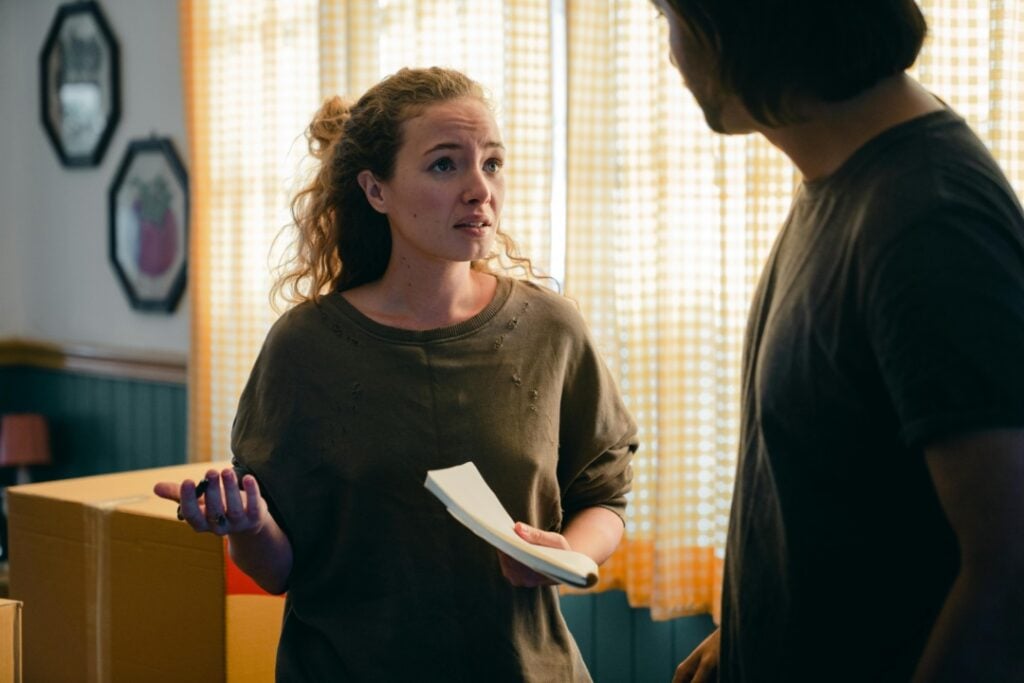 A woman with curly hair holds a notepad and gestures while talking to a man in a room with boxes and checkered curtains.