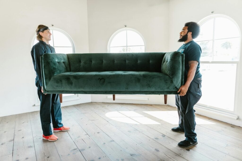 Two people lift a green sofa in a bright room with arched windows and wooden floors.