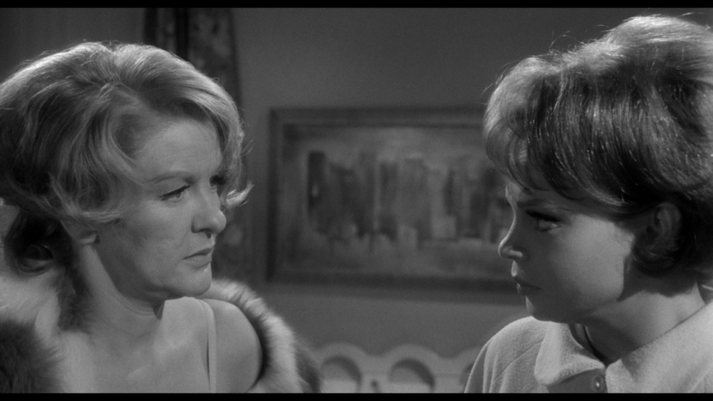 Two women are engaged in a conversation in a black and white setting, with a painting in the background.