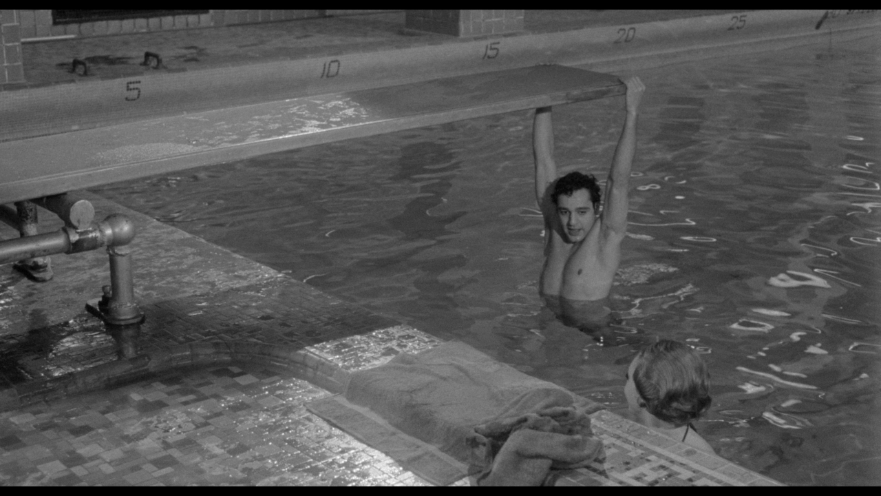 A person hangs from a diving board over a swimming pool, with another person nearby in the water. Towels are on the poolside.