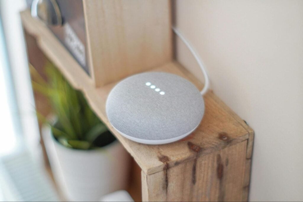A smart speaker with four illuminated dots sits on a wooden shelf next to a potted plant.