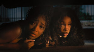Two people with curly hair lie on the ground under a dark object, looking intently to the side.