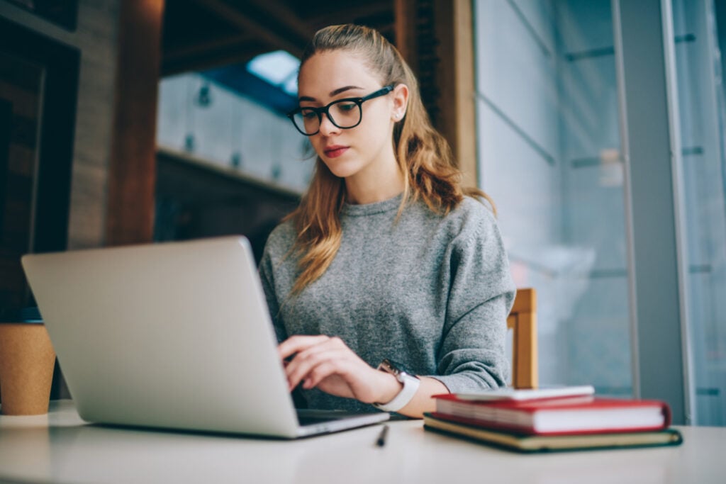A person in glasses works on a laptop at a table with a coffee cup and stacked books nearby.