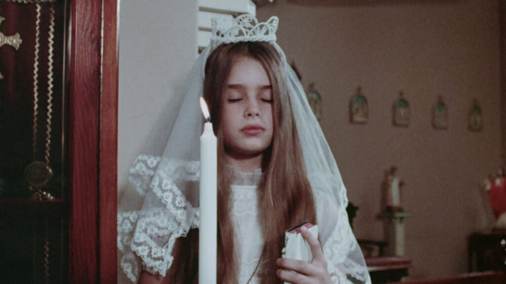 A young girl in a white lace dress and veil stands near a lit candle, eyes closed, holding a small book.