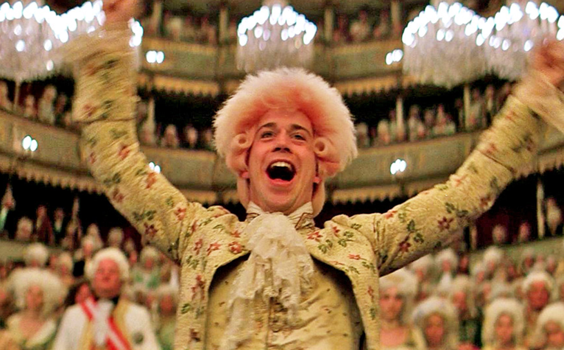 A person in 18th-century costume with a floral pattern and white ruffles, excitedly raising arms in a grand theater setting.