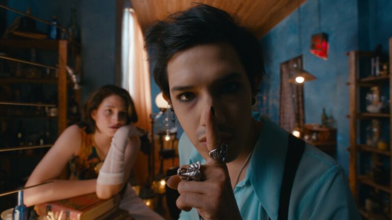 Person holding a finger to their lips, signaling silence, while another person sits in the background. Indoor setting with shelves and a window.