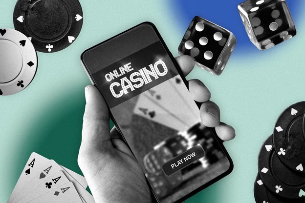 A hand holding a smartphone displaying the text "Online Casino" with a "Play Now" button. Surrounding objects include playing cards, poker chips, and dice on a colorful background.