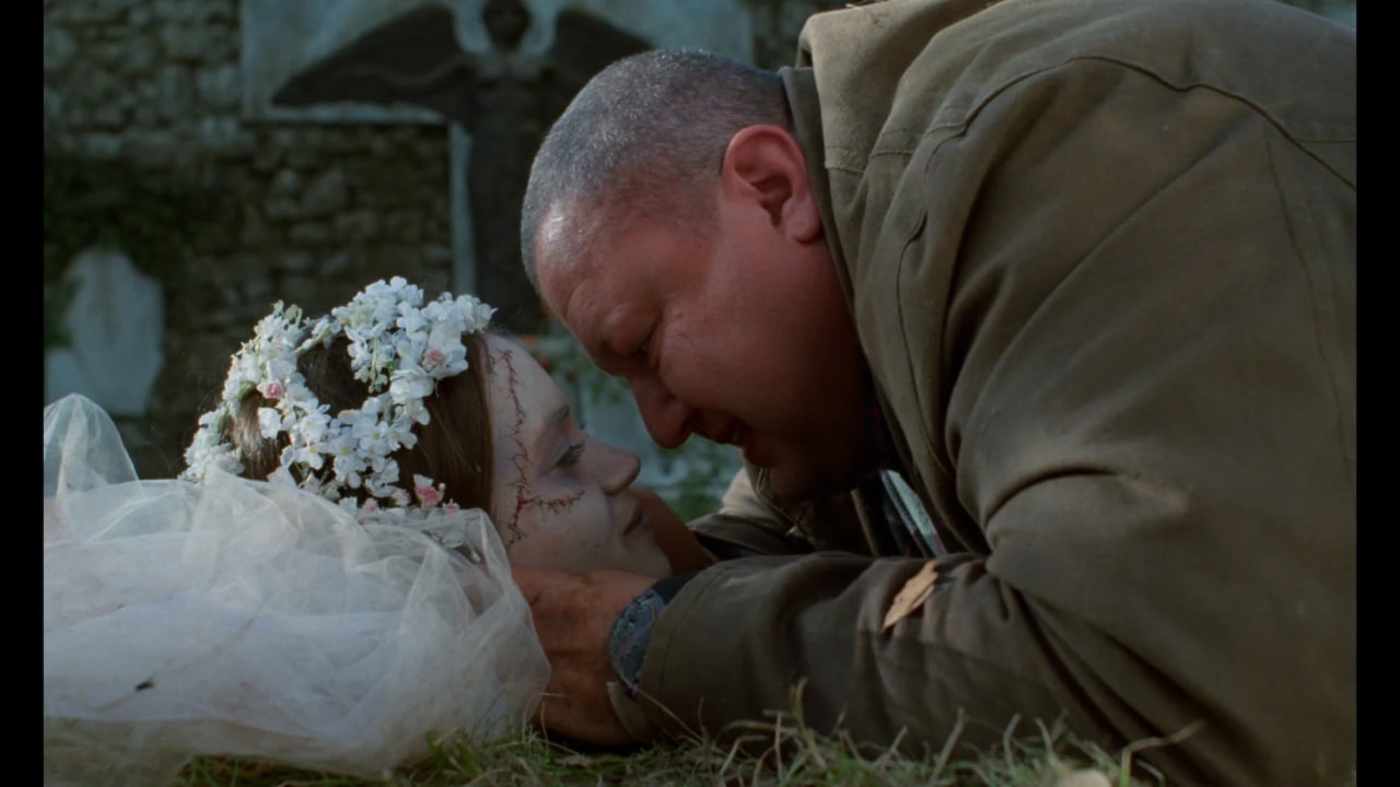 A man in a jacket leans over a woman lying on the ground, wearing a veil and floral headpiece, in a graveyard setting.