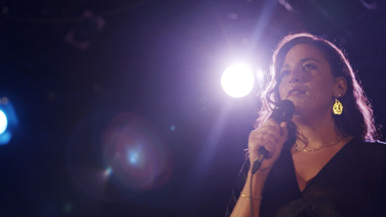 Person standing under bright stage lights, holding a microphone and wearing earrings, mid-performance.