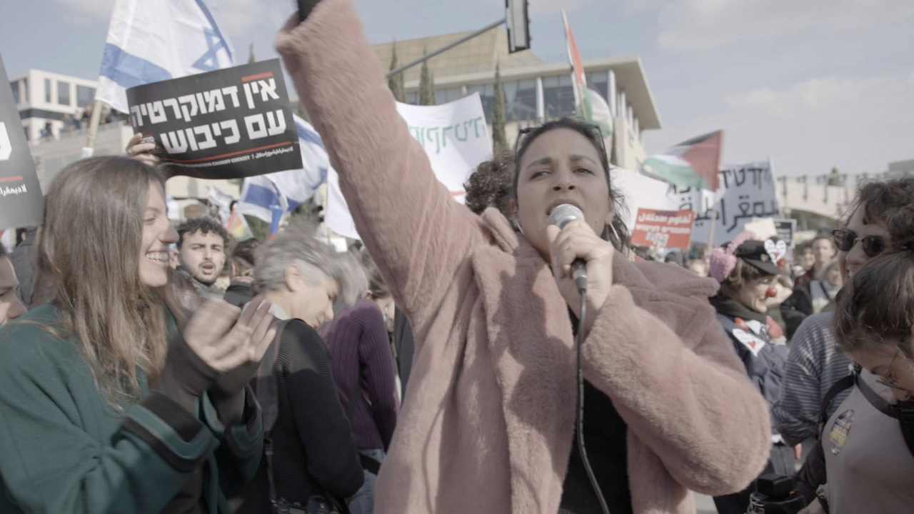 Person speaking into a microphone at a protest, surrounded by people holding signs with various messages.