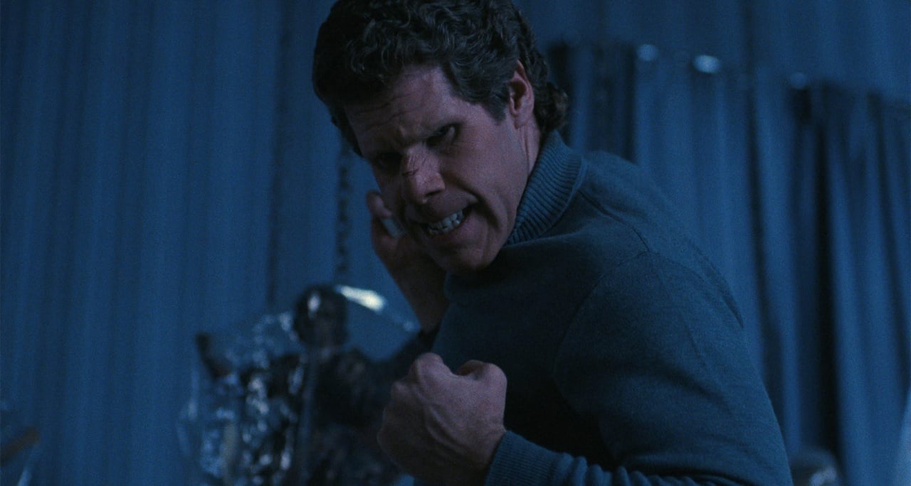 A person in a blue sweater looks intensely to the side, with a tense expression, in a dimly lit room.