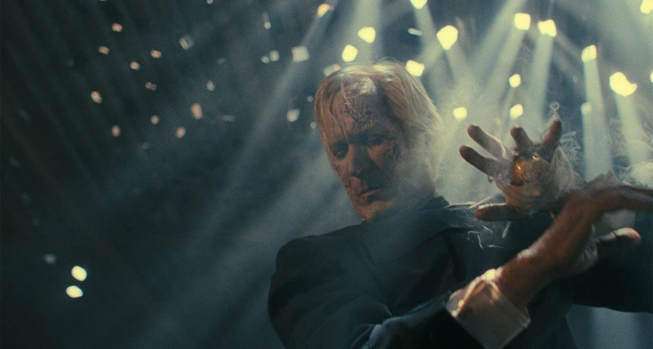 A man in a suit stands under dramatic lighting, with confetti falling around him. His expression is intense, and one hand is raised.