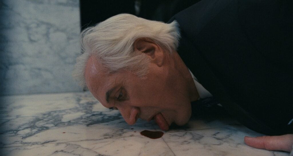 An older man with white hair is on the floor, licking a small puddle of dark liquid on a marble surface.