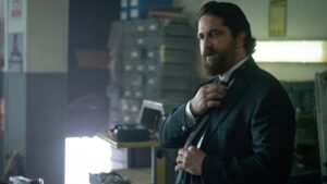 A man in a suit adjusts his bow tie in a dimly lit room with shelves and equipment in the background.
