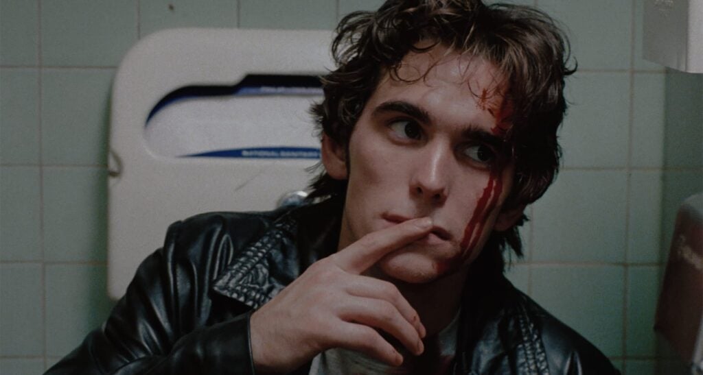 A person with a bloody face wearing a leather jacket sits against a tiled bathroom wall, touching their mouth thoughtfully. A paper towel dispenser is visible in the background.