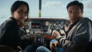 Two people sit in a small airplane cockpit, looking back toward the camera with concerned expressions. The cockpit features various controls and instruments.
