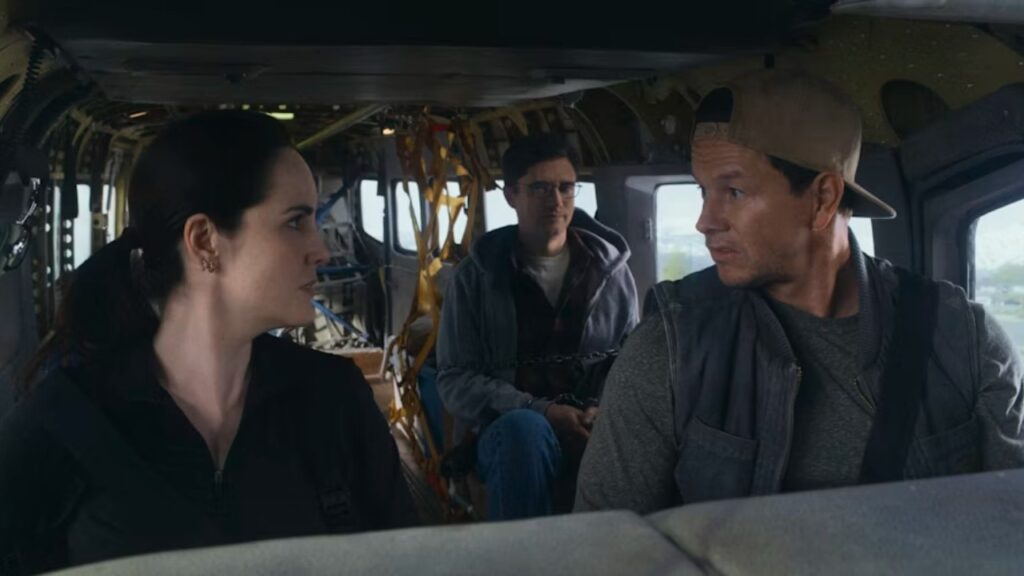 Three people sitting inside a helicopter, two in the front facing left, and one in the back wearing glasses, with chains visible in the background.