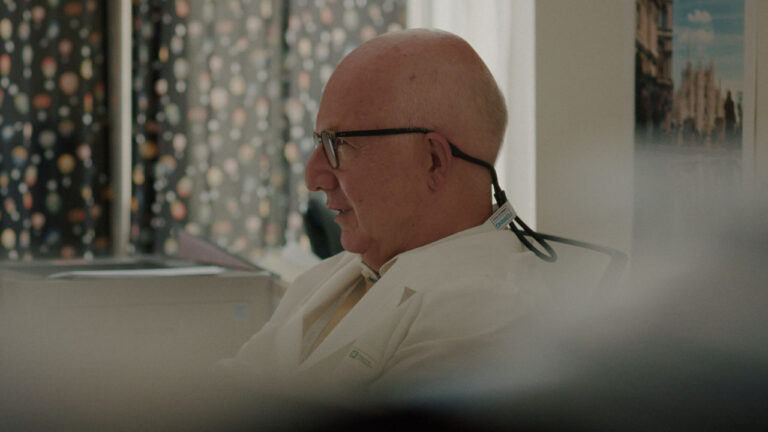 Bald man with glasses and a hearing aid, wearing a white coat, sits sideways in a room with patterned curtains and a poster on the wall.