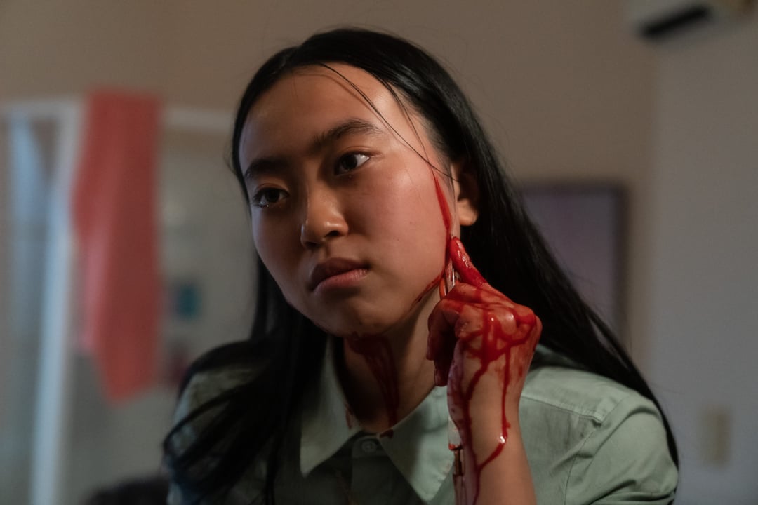 A woman with long hair wipes fake blood from her cheek with her hand, creating streaks. She wears a green shirt and stands indoors with a neutral expression.