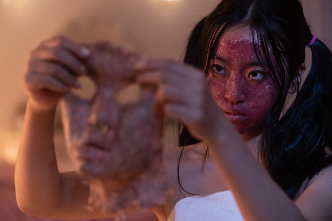 Person with a red, textured face holds up a human-like mask.