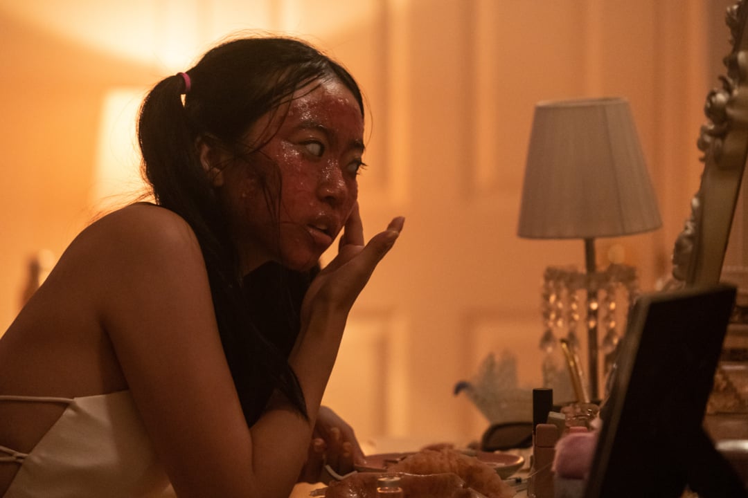 Person with a red facial mask looks in a mirror, touching their cheek. The room has a lamp and various items on the table.