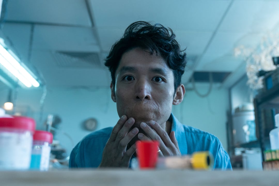 A person in a blue shirt presses their cheeks with their hands while sitting in a room with various jars and containers.
