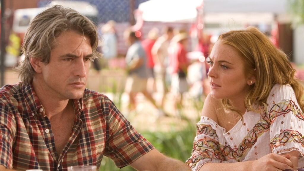 A man and a woman sit at a table outdoors, engaged in conversation. The man wears a plaid shirt and appears thoughtful, while the woman wears a patterned top and looks intently at him.