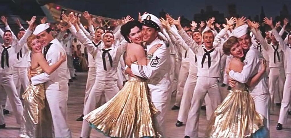 A group of sailors in white uniforms are dancing with women in gold dresses, smiling and raising their arms in celebration on a stage.