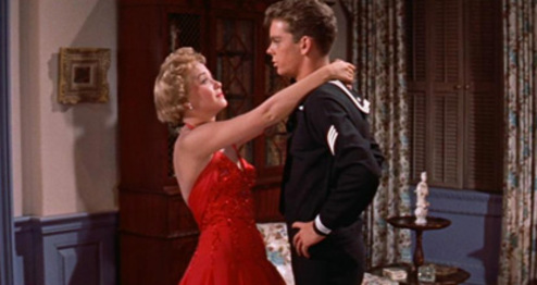 A woman in a red dress embraces a man in a navy uniform, standing in a room with blue walls and a wooden cabinet.