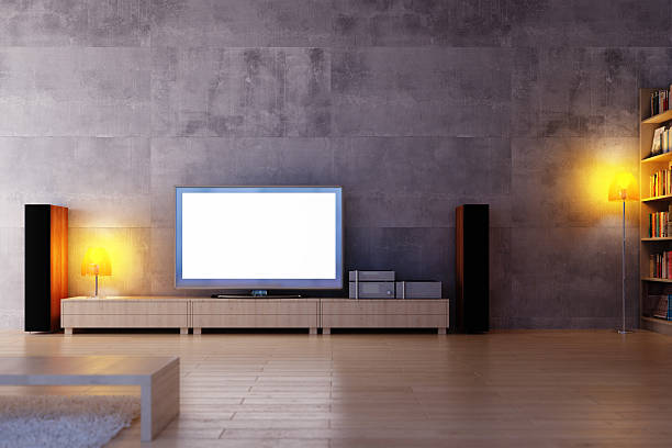 Modern living room with large TV, wooden media console, speakers, and bookshelves. Warm lighting from table lamps adds a cozy ambiance. Minimalist decor and concrete wall.