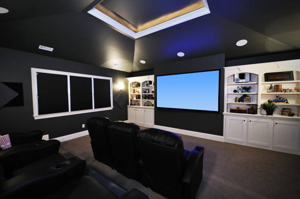 Home theater room with four leather chairs facing a large screen. Shelves with decor and recessed lighting are on the walls, and the ceiling has a tray design.