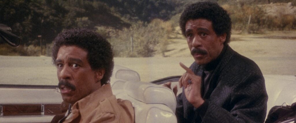 Two men with mustaches sit in a convertible. One wears a beige jacket, looking surprised, the other points outward. Background shows trees and a dirt road.
