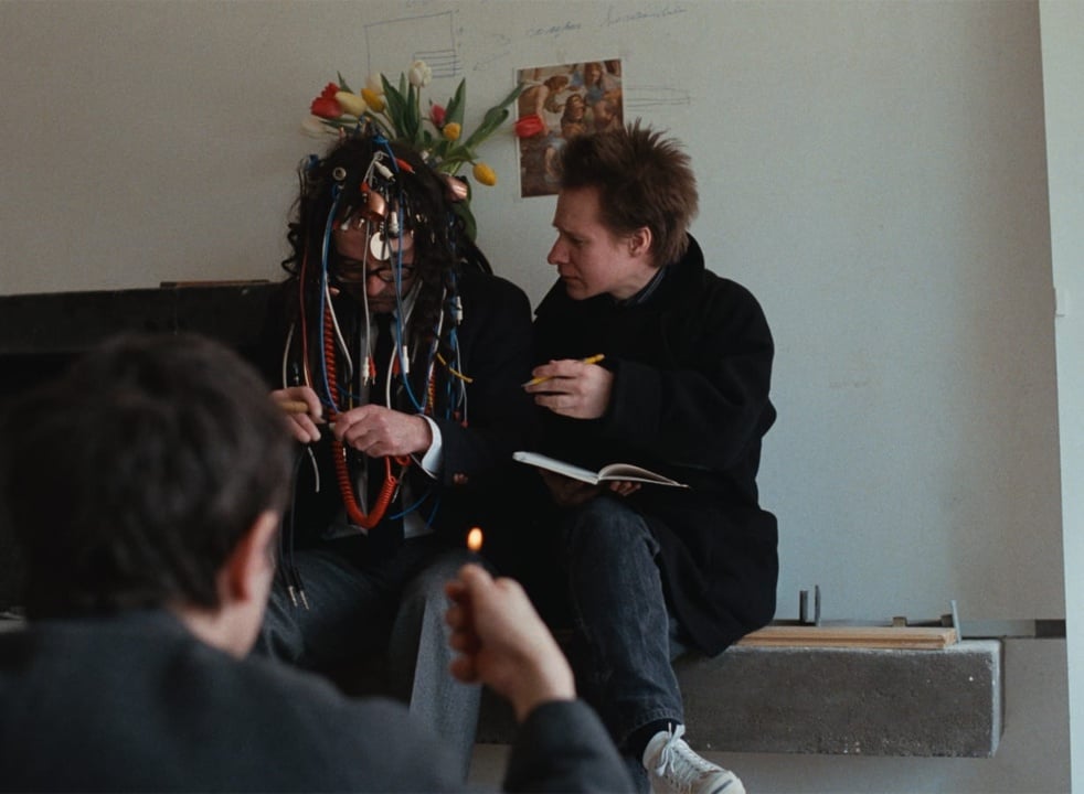 A person with a flower and ribbon headdress is seated next to another holding a pencil and notebook. A third person in the foreground holds up a lighter.