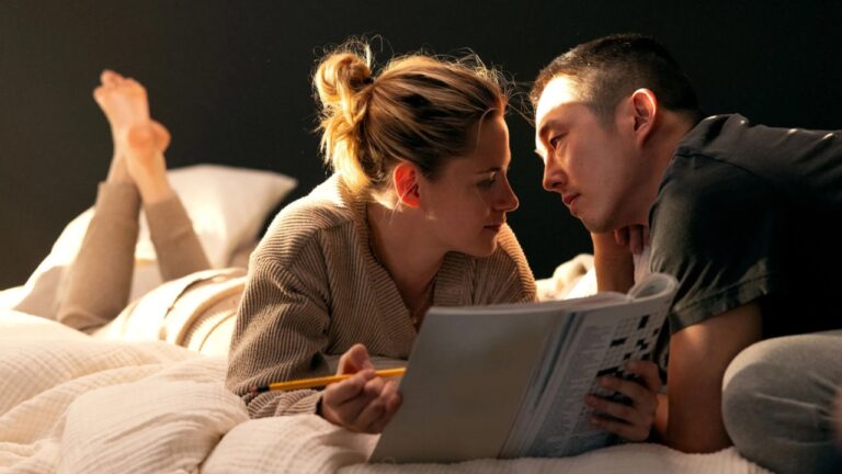 Two people lie on a bed, facing each other, with a book and a pencil in hand.