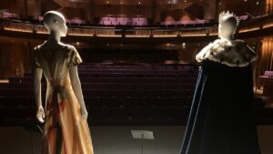 Two mannequins dressed in formal historical costumes stand on a dimly lit stage facing an empty auditorium.