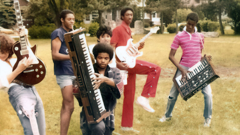 Five people outdoors are posing with musical instruments, including a keyboard, guitars, and a keytar. They appear to be standing on grass with trees and a building in the background.