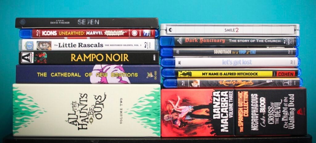 A stack of various DVDs and Blu-rays are arranged on a blue surface against a greenish-blue wall. The titles include films, series, and documentaries in different genres.