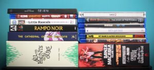A stack of various DVDs and Blu-rays are arranged on a blue surface against a greenish-blue wall. The titles include films, series, and documentaries in different genres.
