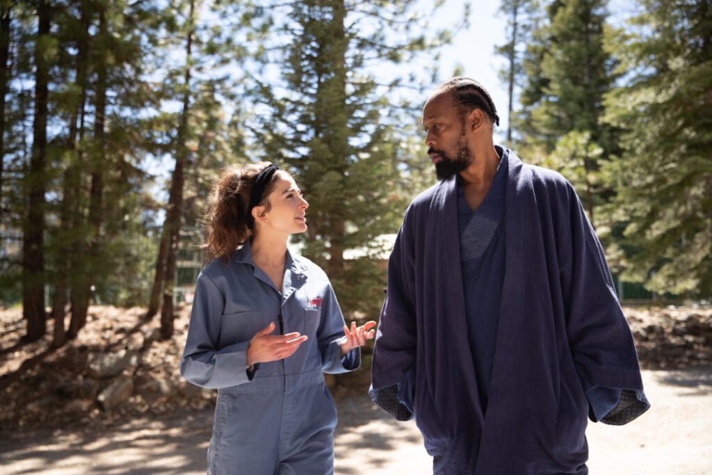 A man and a woman are walking and talking in a wooded area. The woman is wearing a light blue jumpsuit, and the man is wearing a dark robe. Trees are visible in the background.