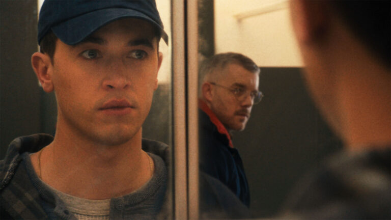 A man in a blue cap with a neutral expression looks at his reflection in a mirror, with another man in glasses blurred in the background.