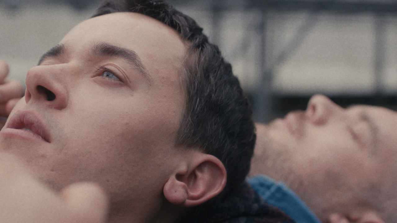 Two individuals lie closely, facing upwards, with thoughtful expressions. One is in the foreground, partially obscured. The background is blurred, featuring indeterminate structures.