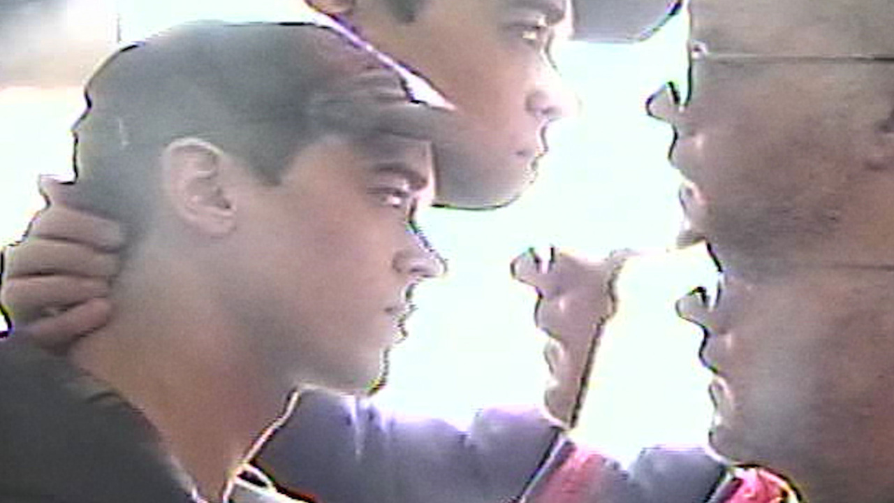 A man is holding another man's face close to his own. The scene is slightly blurry, with strong lighting highlighting their profiles.