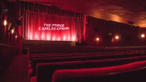 London’s Beloved Prince Charles Cinema Under Threat Of Closure Interior of an empty cinema with red velvet seats and curtains. The screen displays "The Prince Charles Cinema" in white letters.