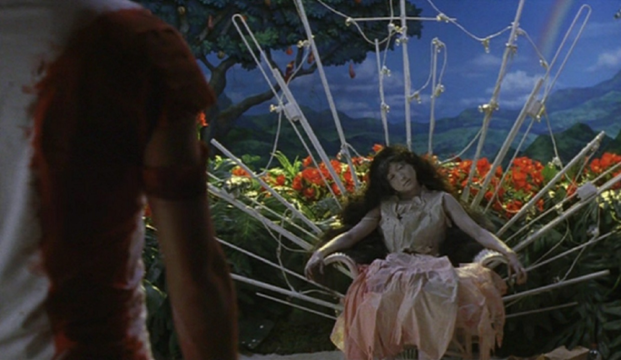 A person sits on a throne-like chair with white pipes, wearing a long dress, surrounded by lush plants and a painted background. Another person stands partially visible in the foreground.