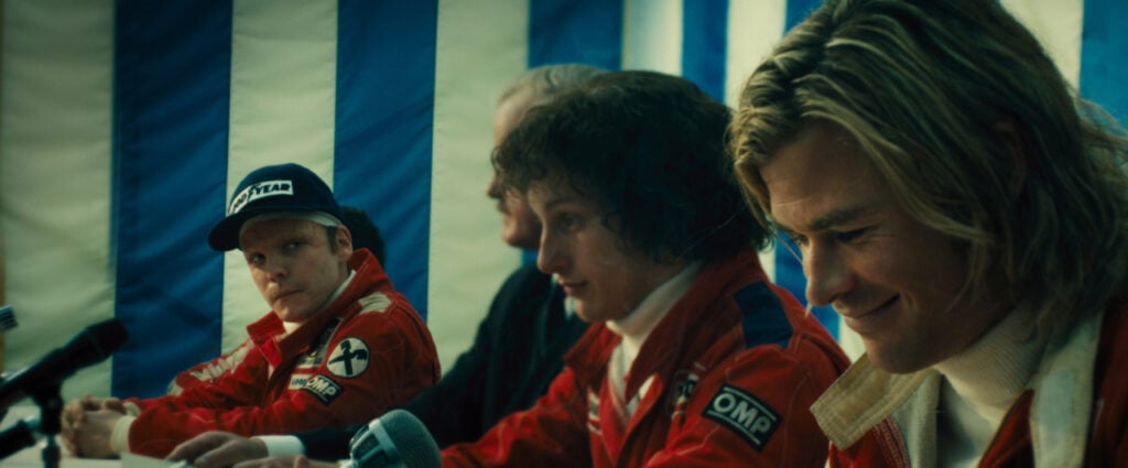 Four men in racing suits sit at a press conference table, microphones in front, with a striped background.