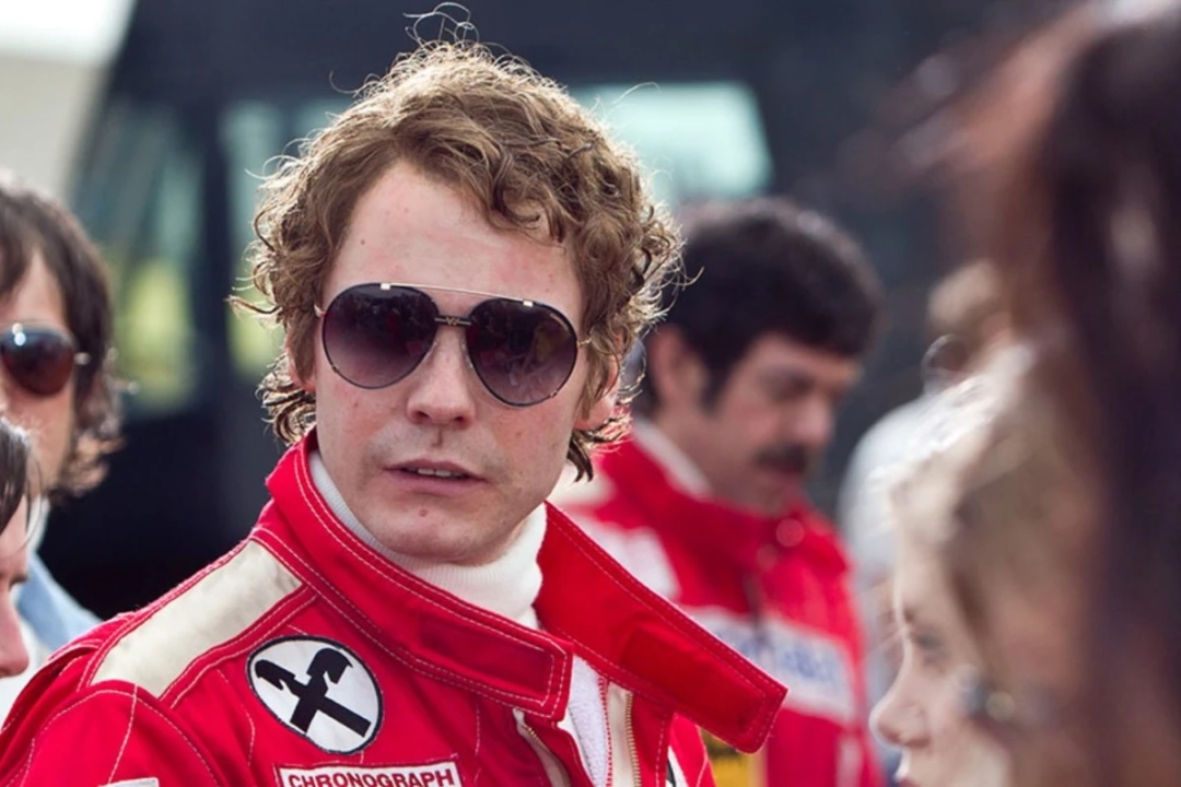 A person in a red racing suit and sunglasses looks to the side, surrounded by others in similar attire.