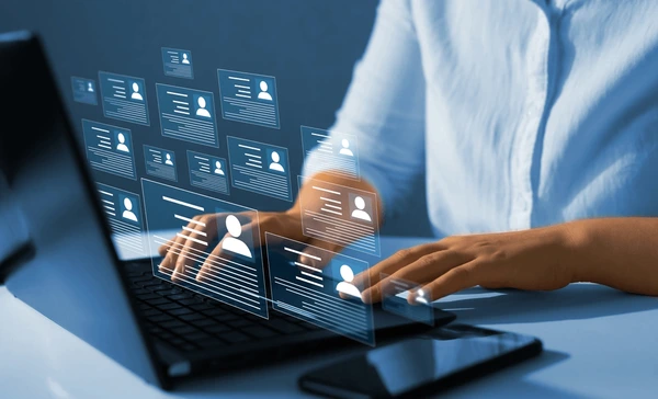 Person typing on a laptop with digital holograms of profiles displayed above the keyboard, next to a smartphone on a desk.