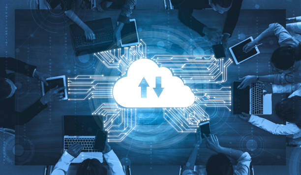 Top view of people using laptops and tablets around a table with a glowing cloud icon featuring upload and download arrows, symbolizing cloud computing and data transfer.
