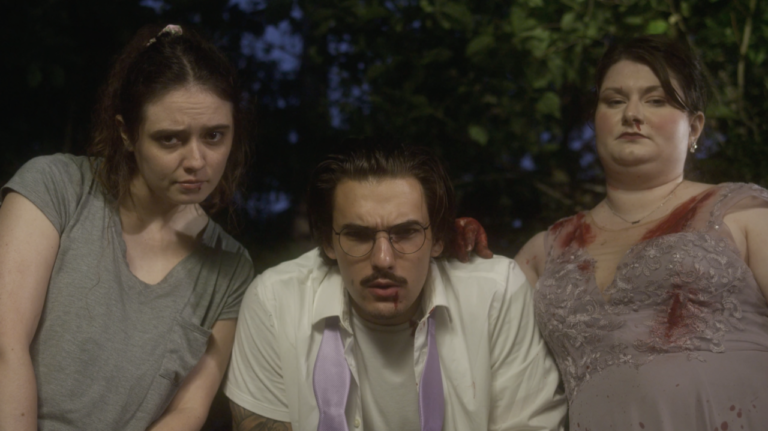 Three people look down at the camera. The man in the middle wears glasses and is flanked by two women. The woman on the right has bloodstains on her dress and chest. They appear serious.