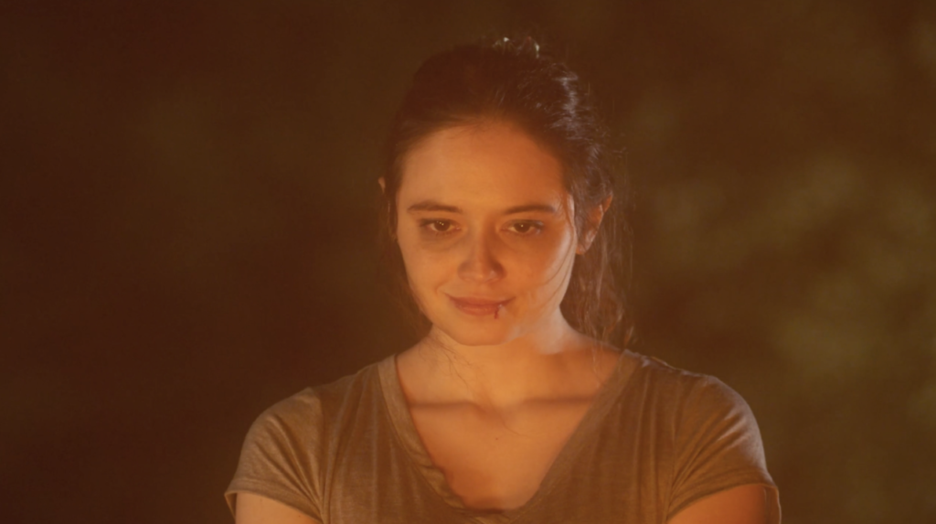 Woman with a neutral expression stands in warm, soft lighting, wearing a gray shirt.